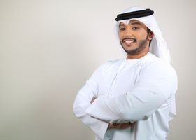 Smiling Middle Eastern man in a white kandura crossing his arms, exuding confidence indoors.
